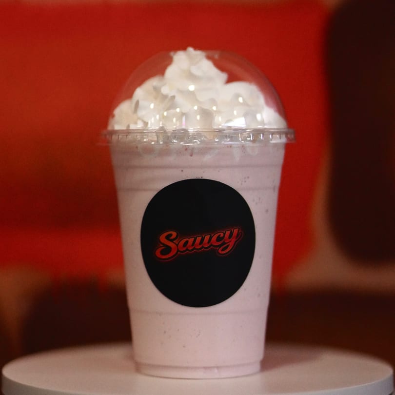 A Saucy branded milkshake with whipped cream on top, against a red and brown background.