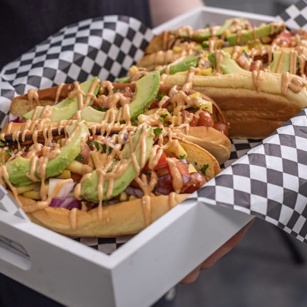 Three gourmet hot dogs topped with avocado, salsa, and sauce, served in a white tray.