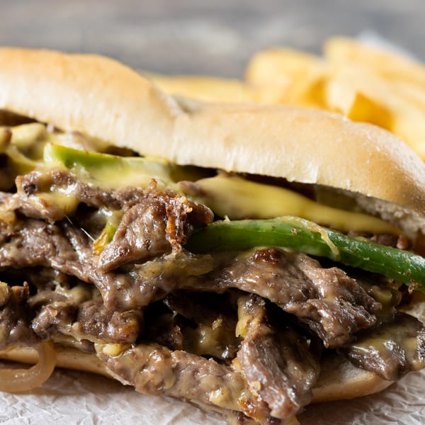 Close-up of a cheesesteak sandwich with melted cheese, green peppers, and steak, with fries in the background.